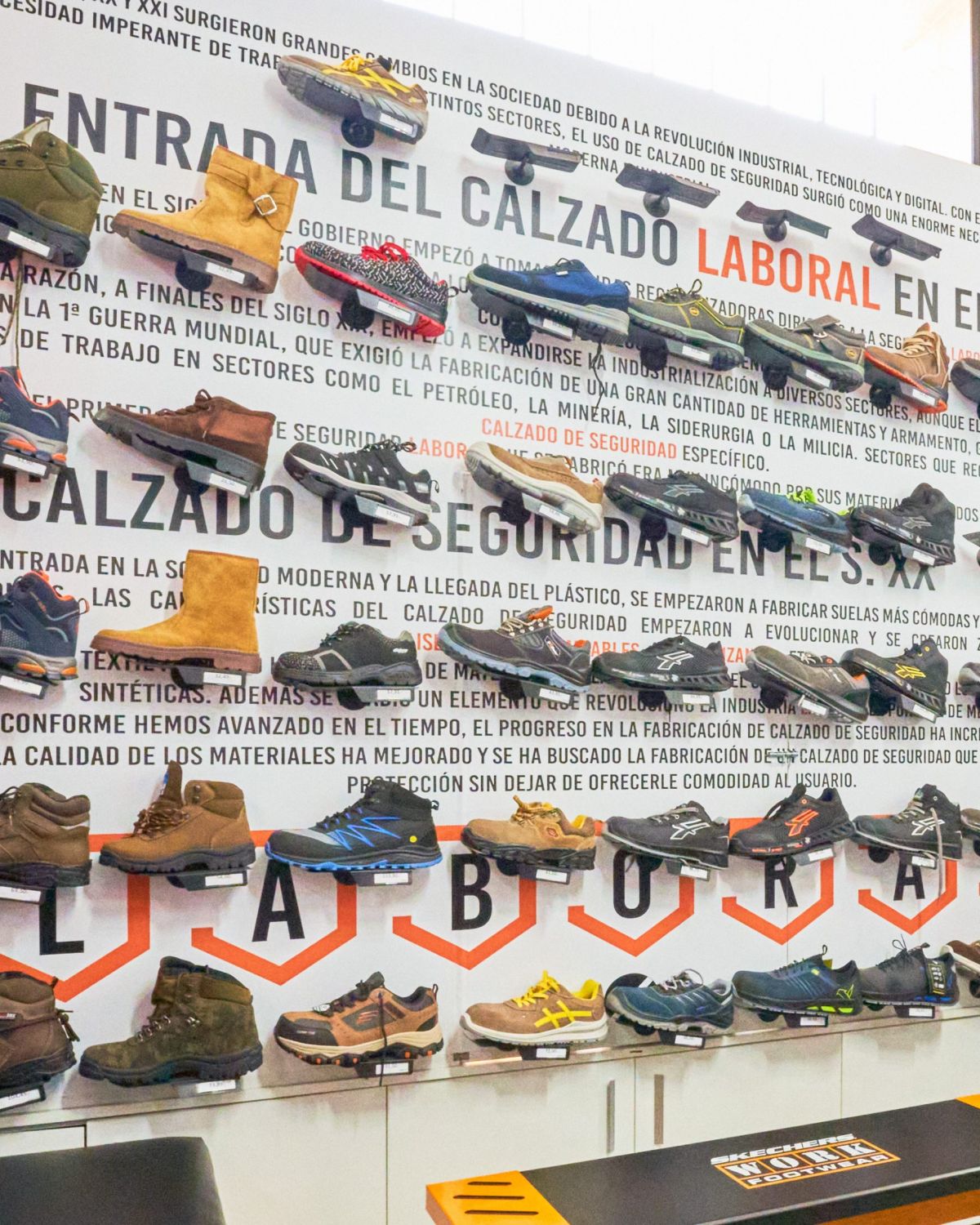Expositor de calzado de seguridad en tienda LABORAL, con variedad de zapatos y botas de protección industrial montados sobre panel informativo sobre la evolución del calzado laboral en España.