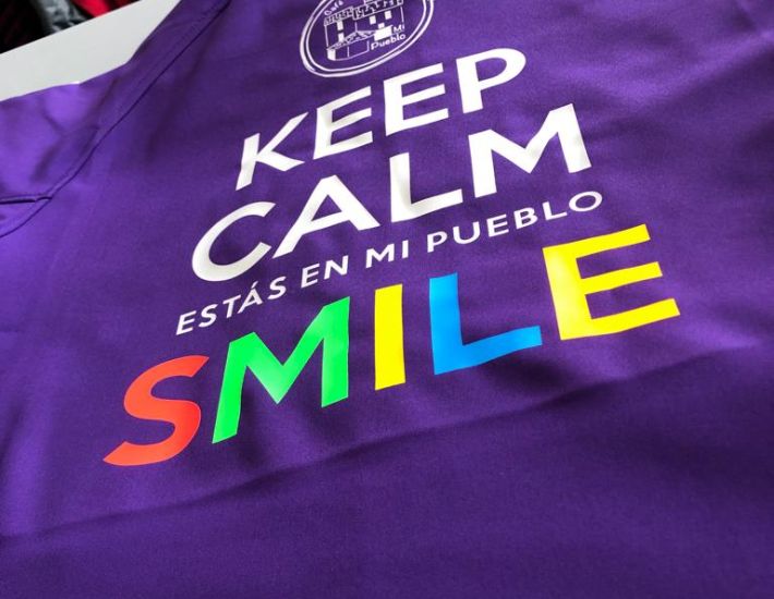 Camiseta deportiva de color morado con el texto “KEEP CALM ESTÁS EN MI PUEBLO SMILE” estampado en blanco, y la palabra “SMILE” en colores del arcoíris. Encima del texto hay un logotipo circular con un dibujo de un bar y las palabras “El Bar de Mi Pueblo”.
