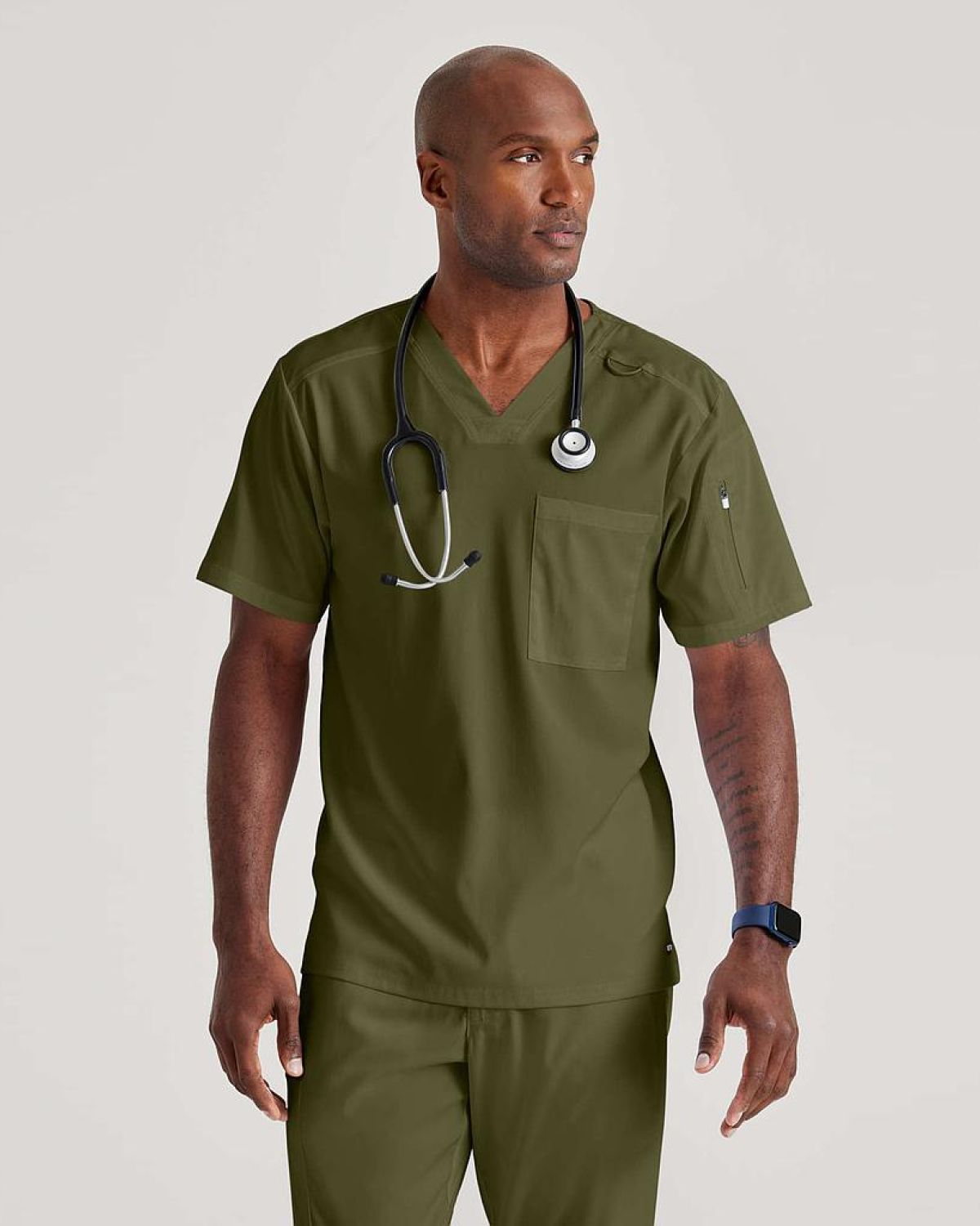 Hombre con uniforme médico verde oliva y estetoscopio, de pie sobre fondo neutro.