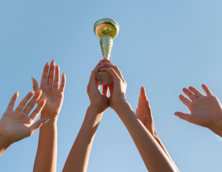 Varias manos alzando un trofeo dorado hacia el cielo, simbolizando celebración, victoria y trabajo en equipo bajo un cielo azul despejado.