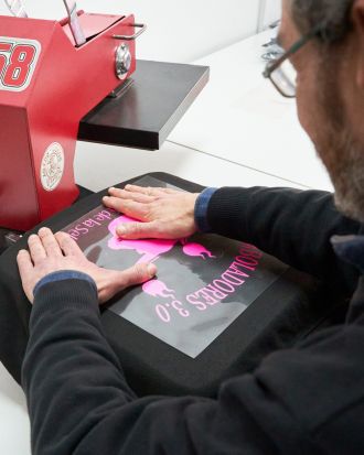 Técnico aplicando un diseño DTF (Direct to Film) en una camiseta negra utilizando una prensa térmica profesional. El gráfico en tonos fucsia muestra el nombre “BOLADORES 3.0” con una figura central, sobre una lámina transparente.