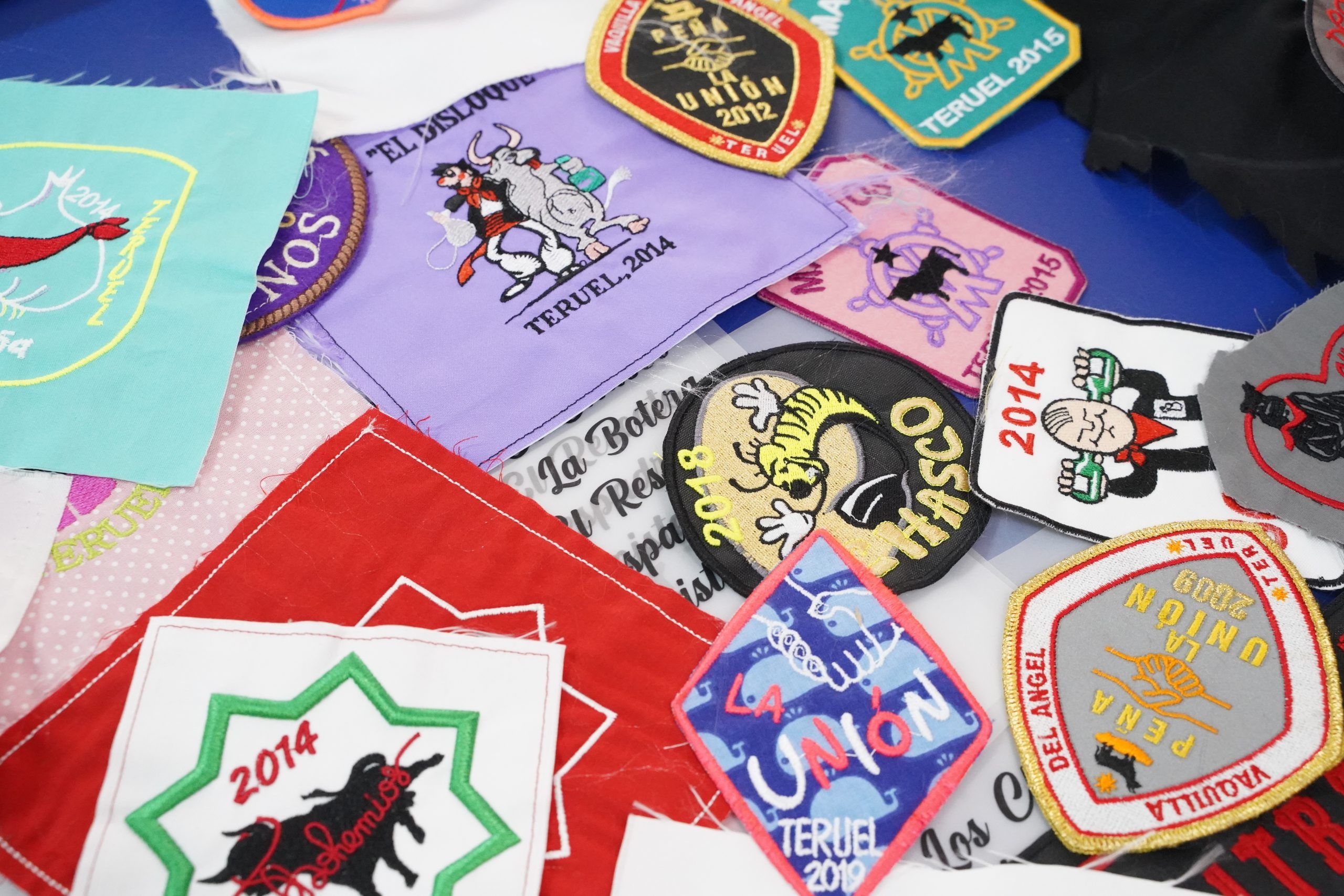 Varios parches bordados coloridos personalizados con nombres de peñas, años y diseños gráficos alusivos a fiestas locales de Teruel, colocados sobre una superficie.