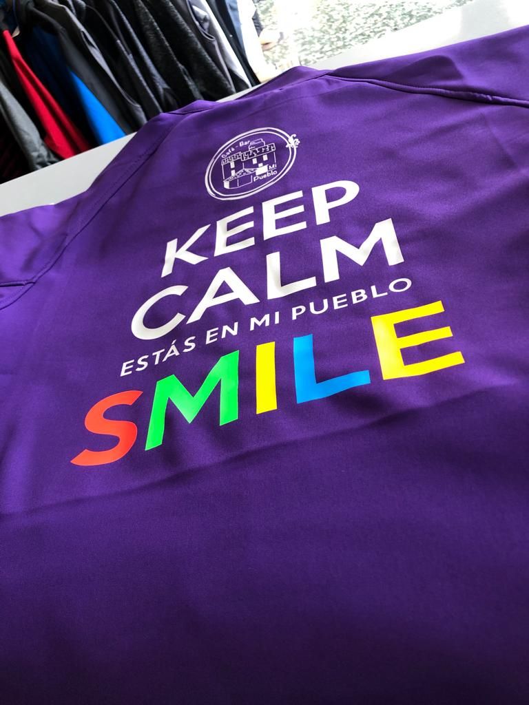 Camiseta deportiva de color morado con el texto “KEEP CALM ESTÁS EN MI PUEBLO SMILE” estampado en blanco, y la palabra “SMILE” en colores del arcoíris. Encima del texto hay un logotipo circular con un dibujo de un bar y las palabras “El Bar de Mi Pueblo”.