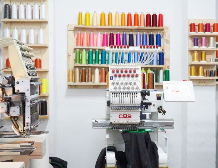 Taller de bordado industrial con máquinas automáticas y estanterías llenas de hilos de colores organizados, listo para personalizar prendas laborales.