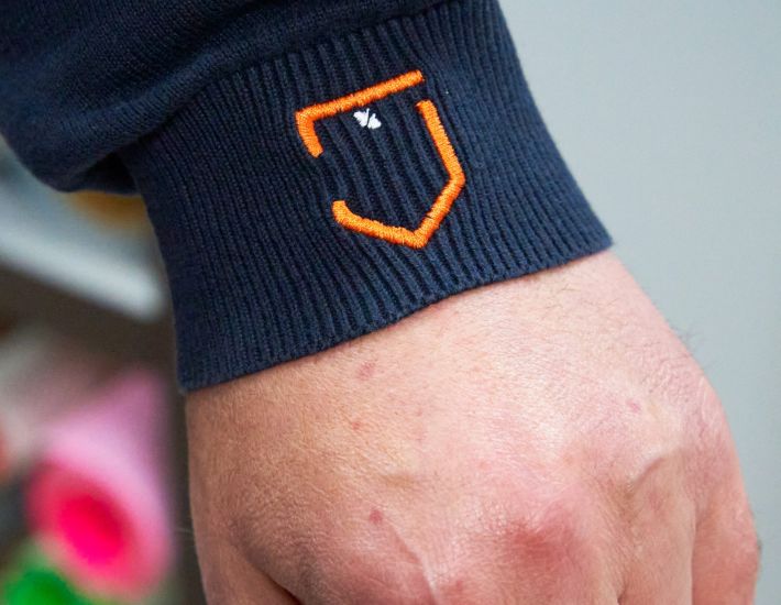 Detalle de un bordado en el puño de una prenda azul marino, con un logotipo en forma de escudo naranja y blanco sobre una mano.