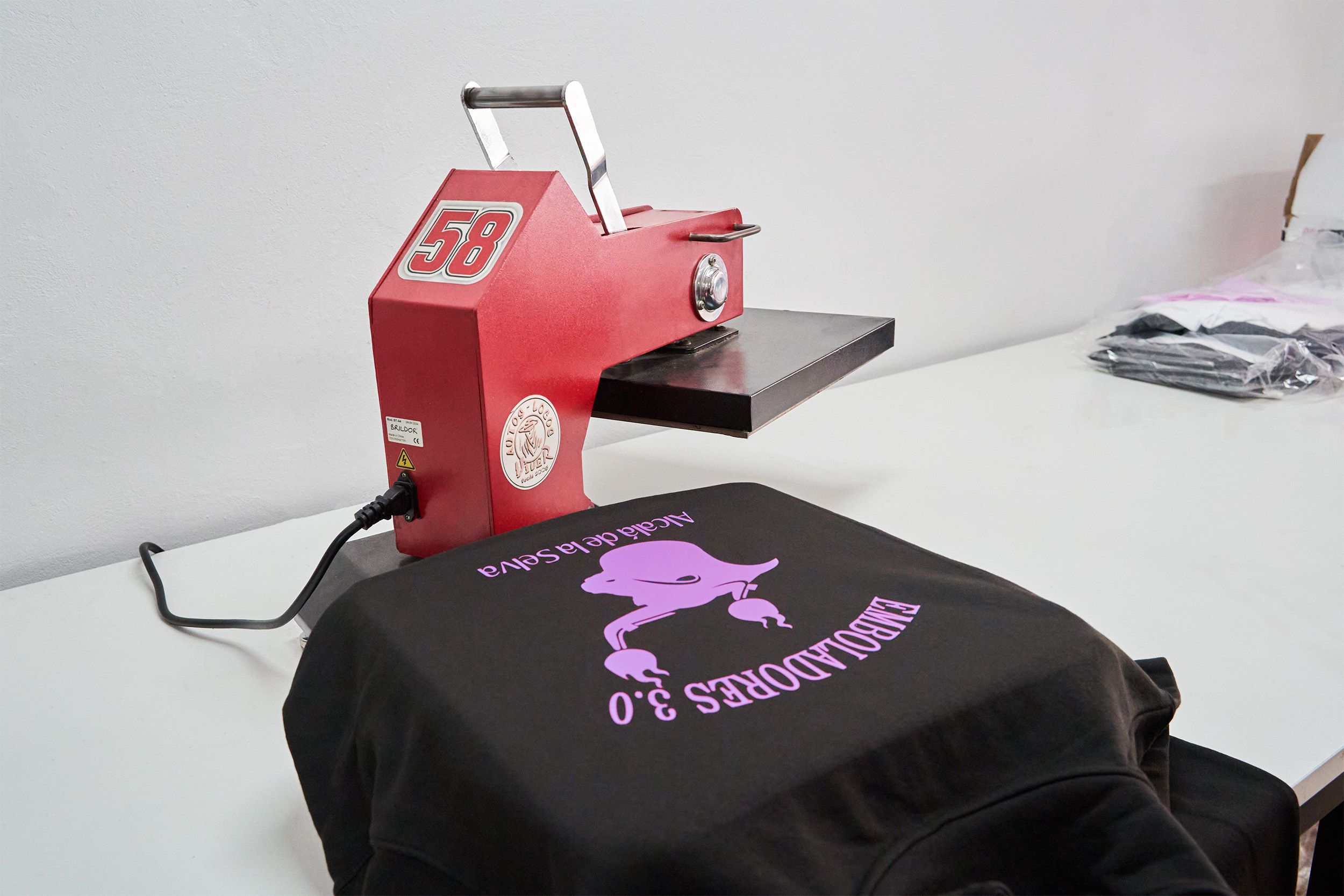 Máquina de serigrafía térmica aplicando diseño morado sobre sudadera negra con texto "EMBOLADORES 3.0" y dibujo de toro.
