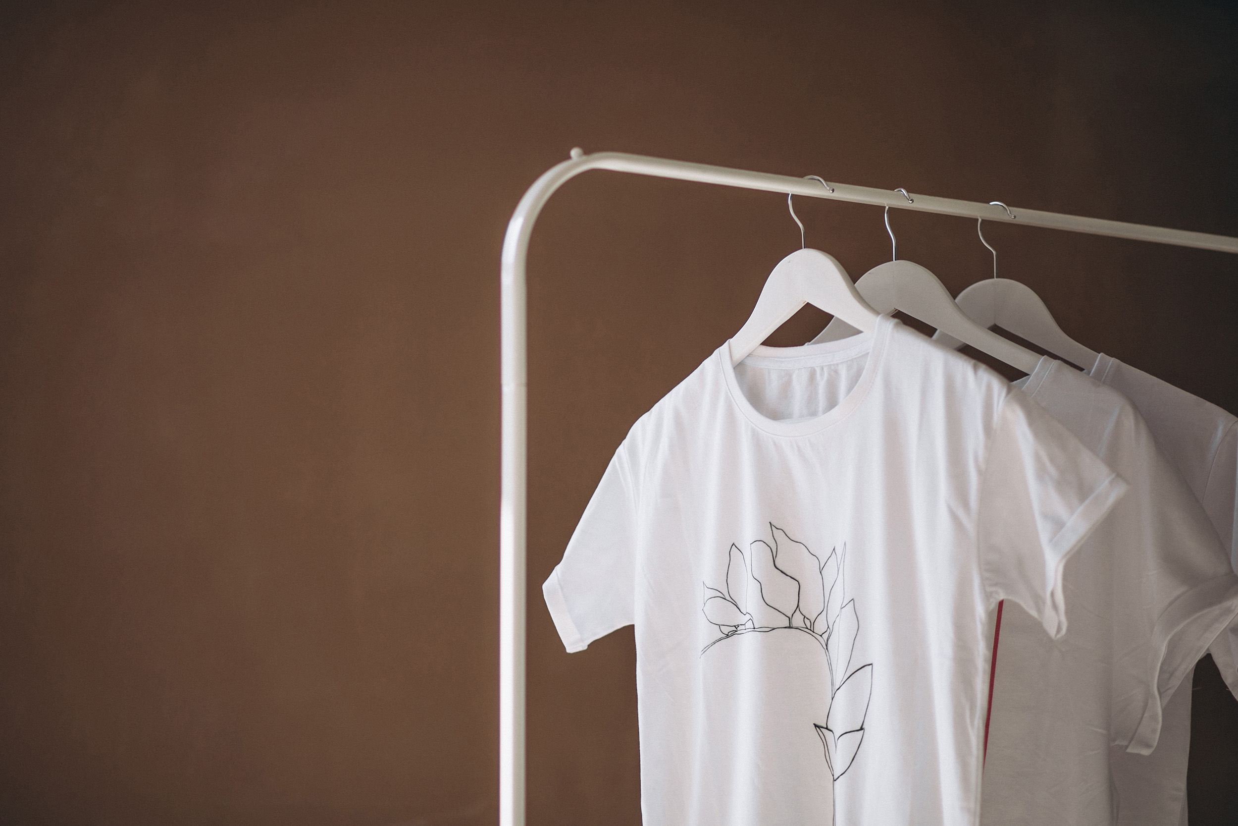 Camisetas blancas colgadas en perchas sobre un perchero blanco, con diseño minimalista de línea de flores en una de ellas, sobre fondo marrón.