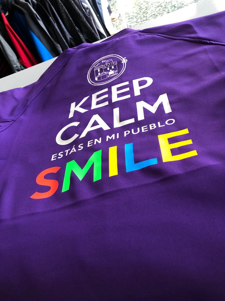 Camiseta deportiva de color morado con el texto “KEEP CALM ESTÁS EN MI PUEBLO SMILE” estampado en blanco, y la palabra “SMILE” en colores del arcoíris. Encima del texto hay un logotipo circular con un dibujo de un bar y las palabras “El Bar de Mi Pueblo”.