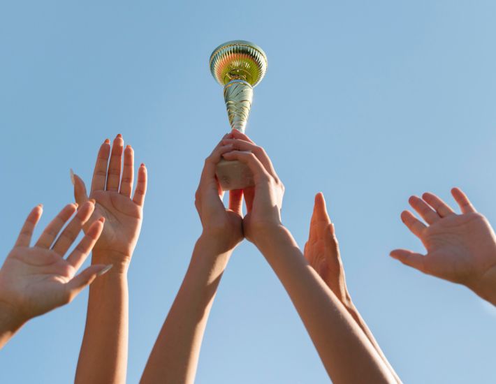 Varias manos alzando un trofeo dorado hacia el cielo, simbolizando celebración, victoria y trabajo en equipo bajo un cielo azul despejado.