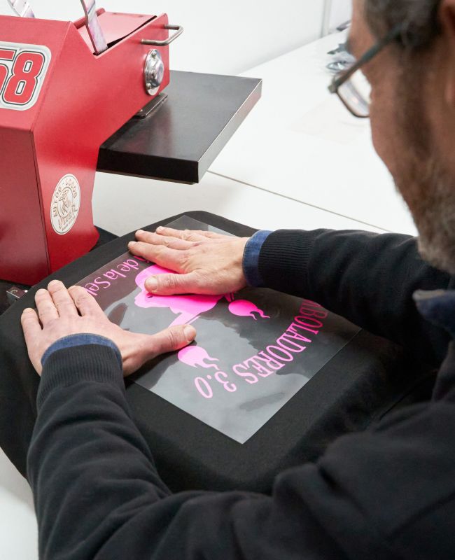 Persona aplicando diseño personalizado sobre camiseta negra con prensa térmica profesional en taller de serigrafía.
