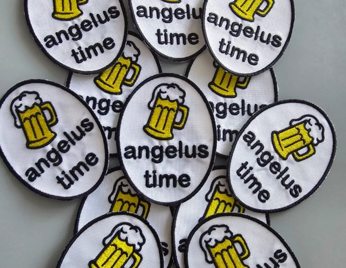 Varios parches bordados ovalados con fondo blanco, borde negro, una jarra de cerveza amarilla espumosa y el texto "angelus time" en negro.