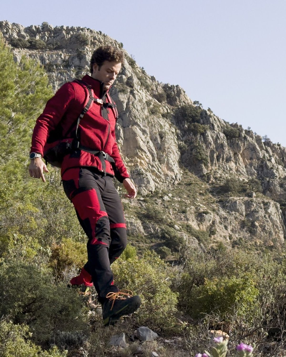 Hombre con ropa de montaña roja y negra caminando por un terreno rocoso y montañoso, rodeado de vegetación y con una mochila a la espalda.