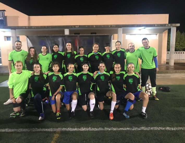 Equipo femenino de fútbol posando en grupo sobre un campo de césped artificial, con uniforme negro y verde fluorescente, acompañado por entrenadores y asistentes.