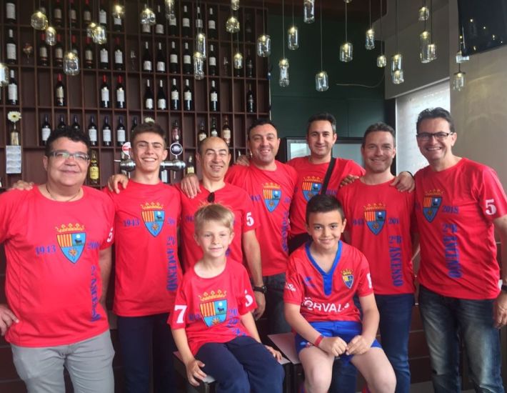 Grupo de hombres y niños con camisetas rojas conmemorativas de un equipo deportivo, posando sonrientes frente a una estantería con botellas de vino en un bar.
