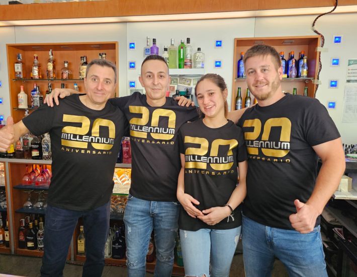 Grupo de cuatro personas posando sonrientes frente a una barra de bar, vestidos con camisetas negras personalizadas con el texto "20 Millennium Aniversario" en letras doradas.