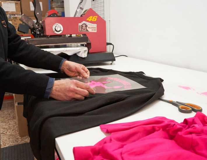 Técnico aplicando un diseño de vinilo rosa sobre una sudadera negra utilizando una plancha térmica profesional. A su alrededor se observan herramientas y materiales textiles, destacando el ambiente de personalización textil de alta calidad.