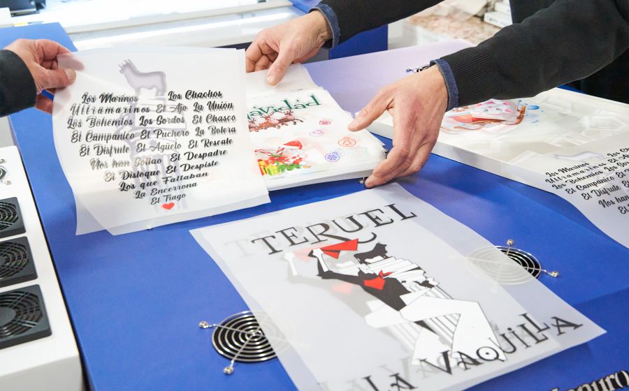 Manos manipulando diseños de serigrafía DTF sobre mesa azul, con motivos festivos y personalizados como "TERUEL LA VAQUILLA" y nombres de peñas, en tienda especializada en estampación textil en Teruel.