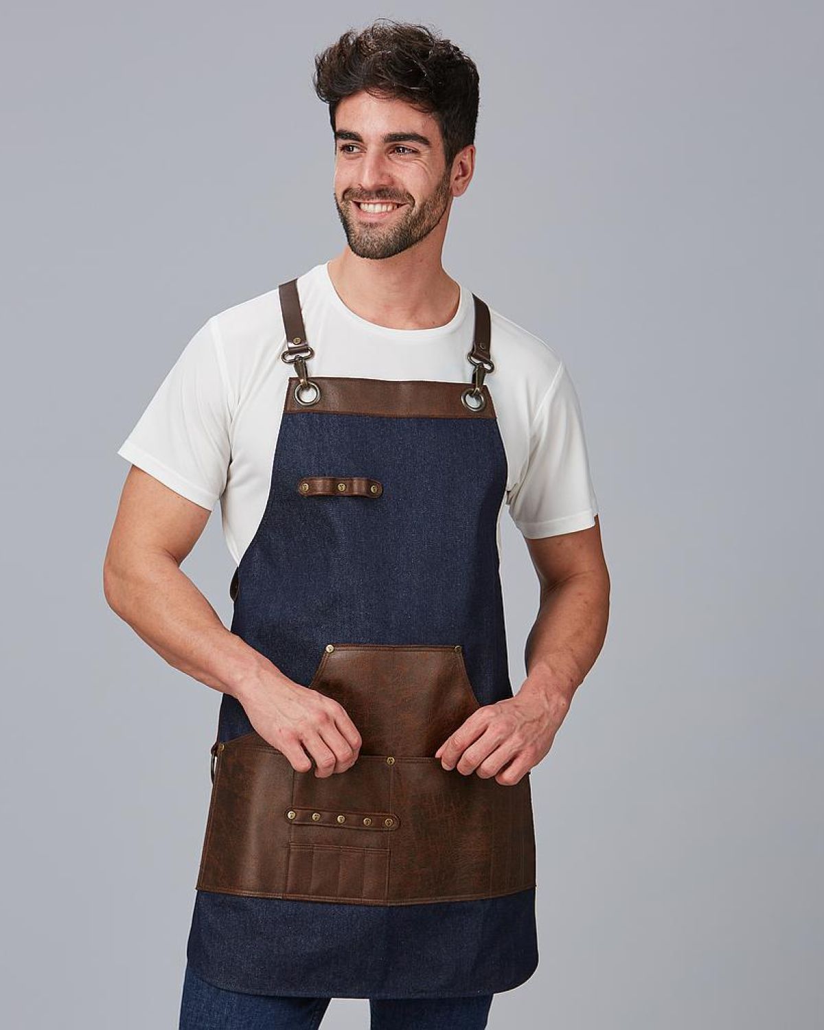 Hombre sonriente con delantal de trabajo estilo barista en mezclilla azul y detalles en cuero marrón, con camiseta blanca y fondo gris neutro.