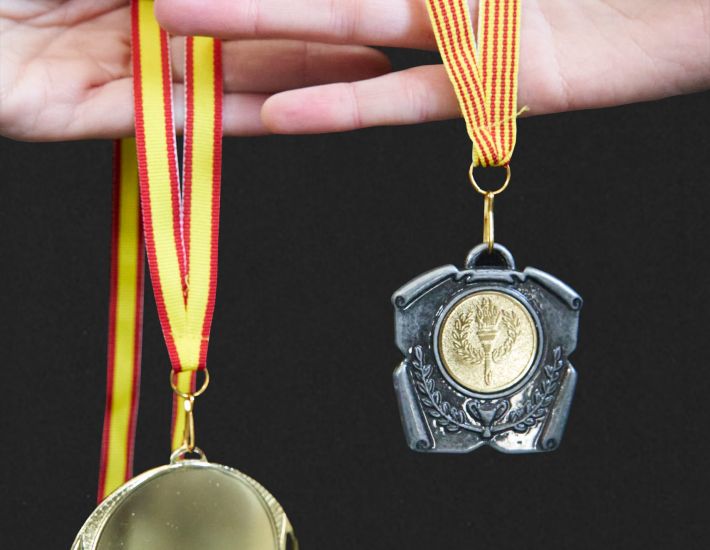 Dos manos sostienen medallas deportivas: a la izquierda, una medalla dorada circular con cinta roja y amarilla; a la derecha, una medalla metálica con forma decorativa y un emblema dorado central de antorcha y laurel, también colgada con cinta roja y amarilla.