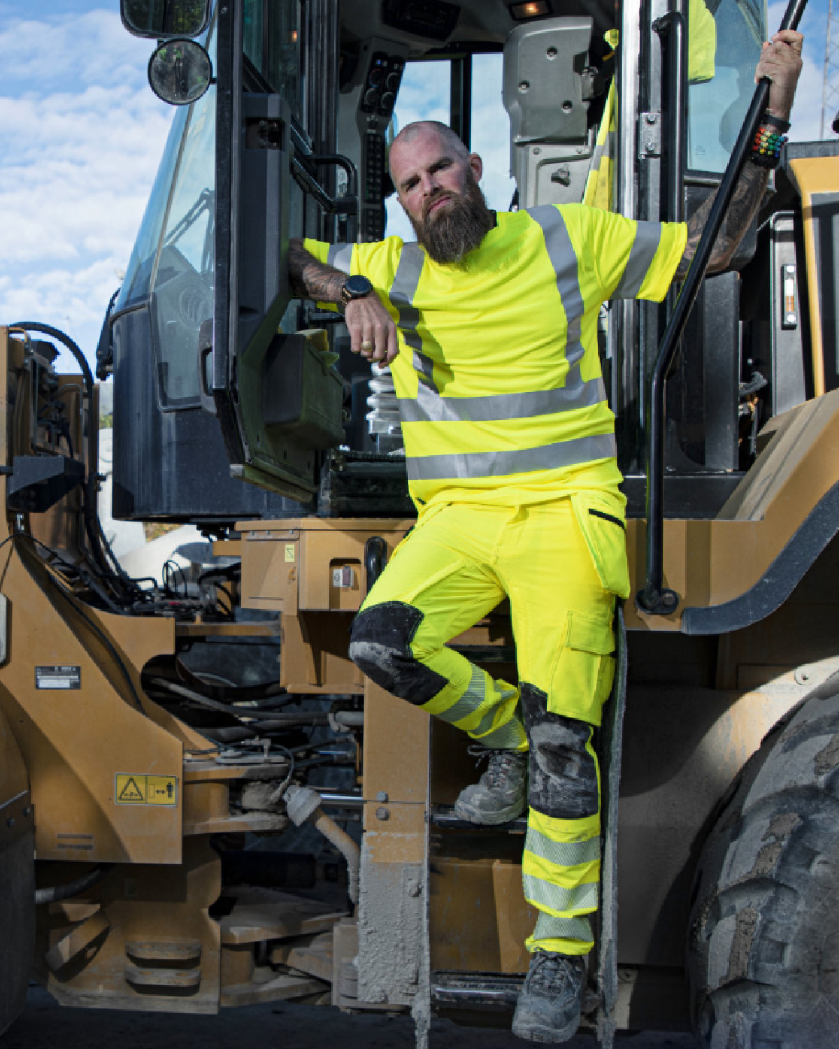 Trabajador con ropa de alta visibilidad amarilla y bandas reflectantes subido a una excavadora en un entorno de obra, con pose relajada.