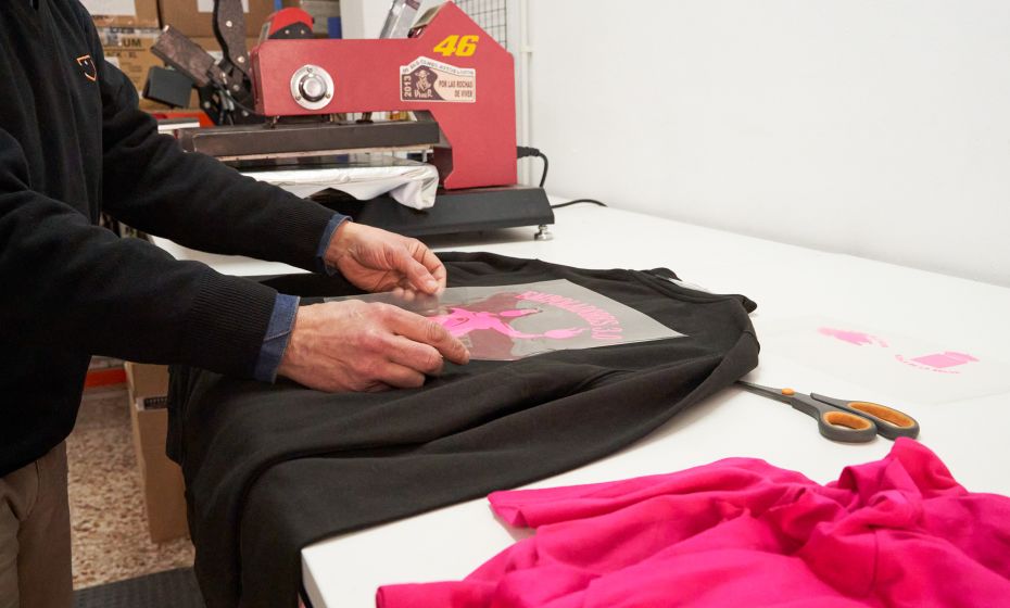 Proceso de estampado textil en taller de serigrafía, aplicando diseño rosa en sudadera negra con plancha térmica, en tienda de ropa laboral en Teruel.