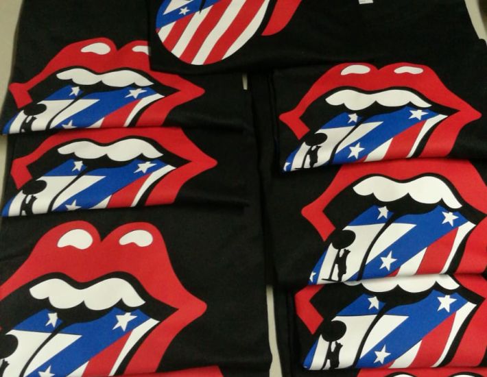 Camisetas negras dobladas con un estampado gráfico grande de unos labios rojos abiertos, mostrando una lengua con los colores y la bandera del Atlético de Madrid. La marca Roly es visible en las etiquetas de las camisetas.