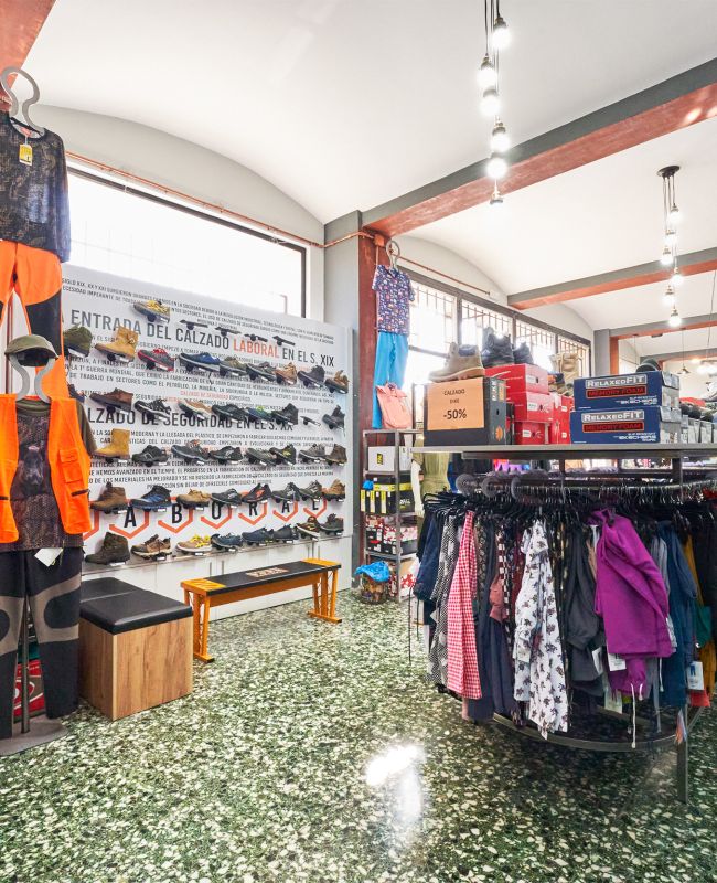 Vista interior de tienda LABORAL con ropa y calzado de trabajo, expositores con chaquetas y camisas, zona de calzado de seguridad y señal de promoción del 50%.