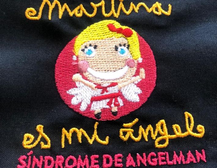 Bordado en tela negra con dibujo de una niña sonriente con alas y texto "Martina es mi ángel. Síndrome de Angelman"