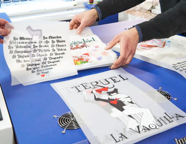 Dos personas manipulan hojas de transfer DTF sobre una mesa azul, con diseños coloridos y textos como "TERUEL", "LA VAQUILLA" y nombres de peñas. Muestra el proceso de personalización textil.