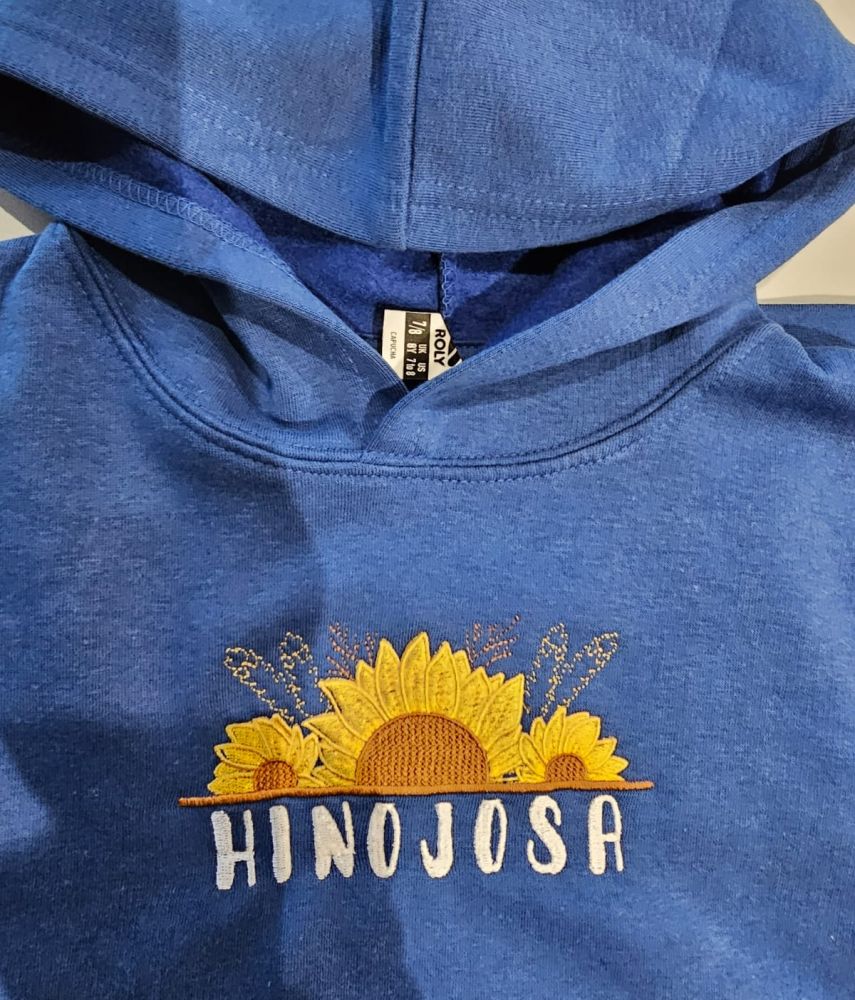 Sudadera azul con capucha de la marca Roly, bordada con girasoles amarillos y la palabra "Hinojosa" en letras blancas.