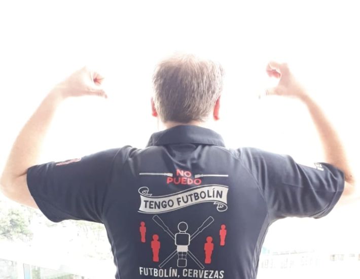 Hombre de espaldas levantando los brazos con una camiseta negra personalizada que dice: "No puedo, tengo futbolín. Futbolín, cervezas y cubatas. En ese orden", junto a gráficos de figuras de futbolín.